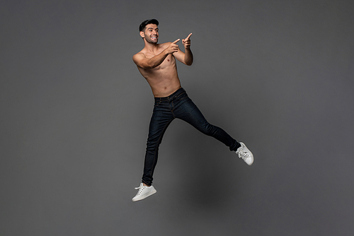 Handsome shirtless smiling Caucasian man jumping and pointing hands to copy space aside in gray isolated studio background