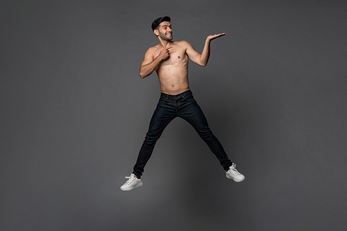 Handsome shirtless smiling Caucasian man jumping with hand opening to empty space aside in gray isolated studio background