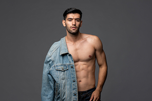 Young handsome sexy half shirtless man with denim jacket posing in light gray isolated studio background