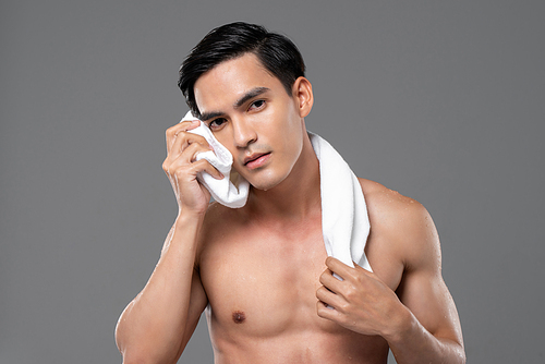 Beauty and skin care portrait of shirtless handsome young Asian man wiping his face with towel isolated on gray studio background