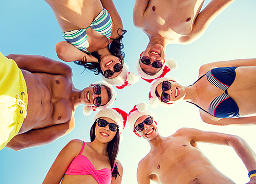 friendship, christmas, summer vacation, holidays and people concept - group of smiling friends wearing swimwear and santa helper hats standing in circle over blue sky