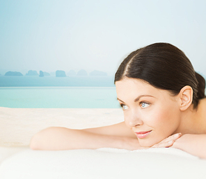 people, beauty and body care concept - happy beautiful woman lying on massage desk at spa resort over sea and infinity pool background