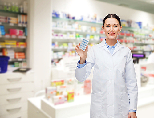 medicine, pharmacy, people, health care and pharmacology concept - happy young woman pharmacist with pills over drugstore background