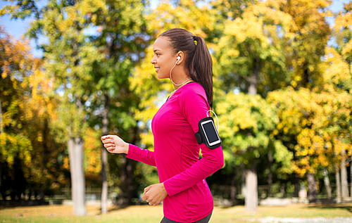 sport, fitness, technology and people concept - smiling young african american woman running with smartphone and earphones over autumn park background