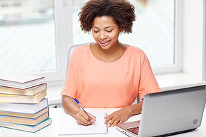 people, technology and education concept - happy african american young woman sitting at table with laptop computer and books at home