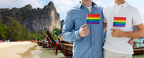 people, homosexuality, same-sex marriage, travel and love concept - close up of happy male gay couple holding rainbow flags and hugging over thailand beach background