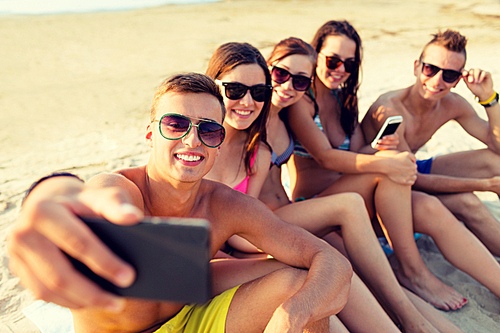 friendship, leisure, summer, technology and people concept - friends sitting and taking selfie with smartphone on beach