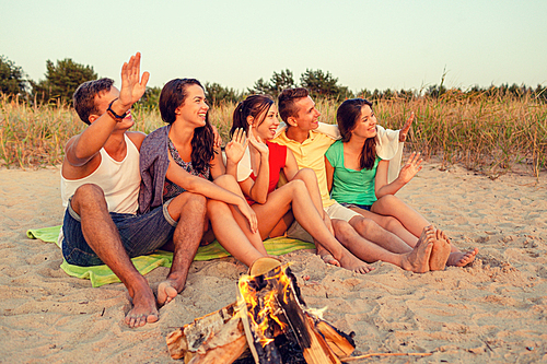 friendship, summer vacation, holidays, gesture and people concept - group of smiling friends sitting near fire and waving hands on beach