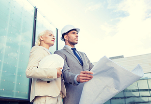 business, partnership, architecture and people concept - businessman and businesswoman with blueprint and helmets on city street