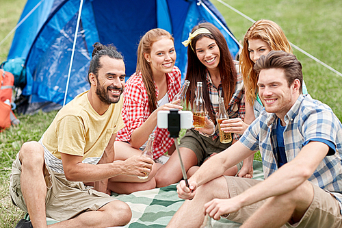 camping, travel, tourism, hike and people concept - happy friends with glass bottles drinking cider or beer and taking picture by smartphone on selfie stick at camping