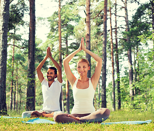 sport, fitness, yoga and people concept - smiling couple meditating and sitting on mats with raised hands over green woods background