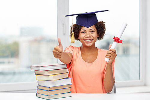 education, school, graduation, gesture and people concept - happy smiling african american student girl in bachelor cap with books and diploma sitting at table and showing thumbs up at home