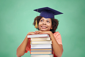 education, school, knowledge and people concept - happy smiling african american student girl in bachelor cap with books sitting at table and dreaming over green chalk board background