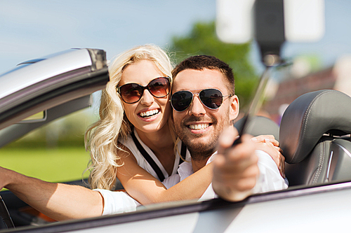 road trip, leisure, couple, technology and people concept - happy man and woman driving in cabriolet car and taking picture with smartphone on selfie stick