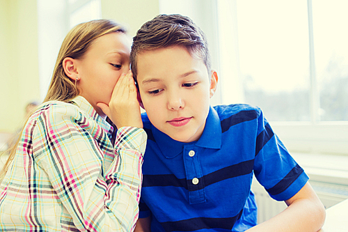 education, elementary school, learning and people concept - smiling schoolgirl whispering secret to classmate ear in classroom