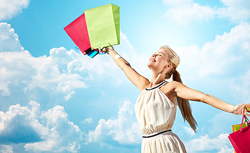consumerism, sale and people concept - smiling woman with shopping bag rising hands over blue sky and clouds background