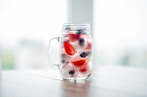 healthy eating, drinks, weight loss and detox concept - close up of fruit water with strawberry, blackcurrant or blueberry and ice cubes in glass mug on table