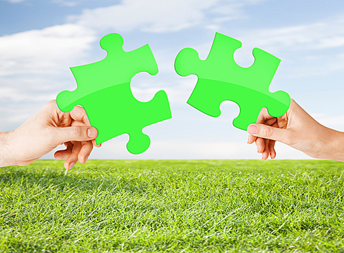 nature, ecology, energy saving, people and environment concept - close up of couple hands trying to connect green puzzle pieces over blue sky and grass background