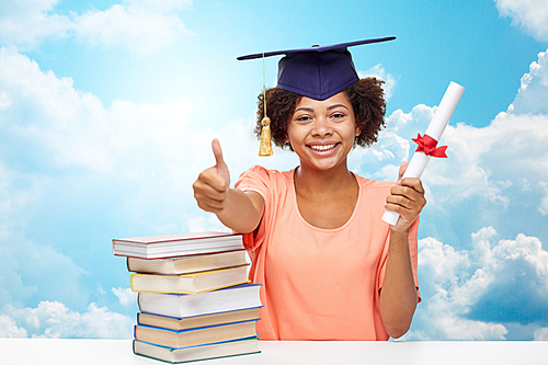 education, school, graduation, gesture and people concept - happy smiling african american student girl in bachelor cap with books and diploma showing thumbs up over blue sky and clouds background