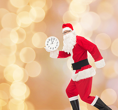 christmas, holidays and people concept - man in costume of santa claus running with clock showing twelve over beige lights background