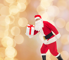 christmas, holidays and people concept - man in costume of santa claus running with gift box over beige lights background