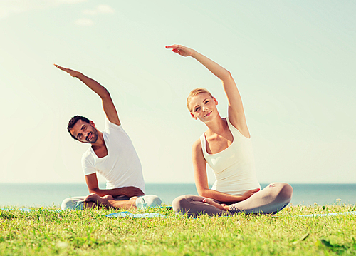 fitness, sport, friendship and lifestyle concept - smiling couple making yoga exercises sitting on mats outdoors