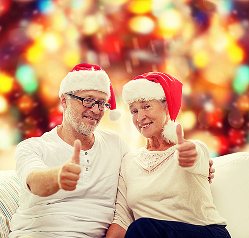 family, holidays, christmas, age and people concept - happy senior couple in santa helper hats sitting on sofa over red lights background and showing thumbs up gesture