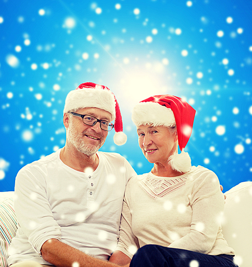 family, holidays, christmas, age and people concept - happy senior couple in santa helper hats sitting on sofa over blue snowy background