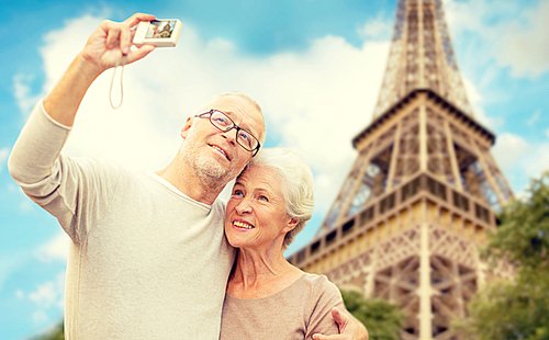 age, tourism, travel, technology and people concept - senior couple with camera taking selfie on street over eiffel tower background