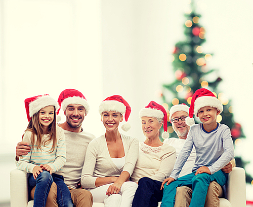 family, happiness, generation, holidays and people concept - happy family in santa helper hats sitting on couch over living room and christmas tree lights background