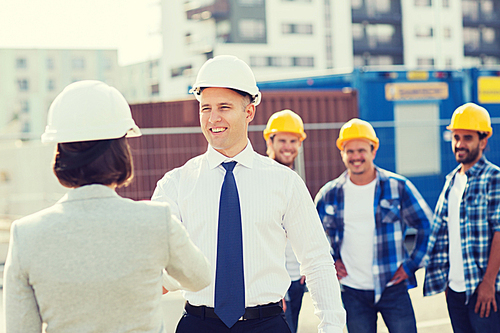 business, building, teamwork, gesture and people concept - group of smiling builders in hardhats with clipboard greeting each other outdoors