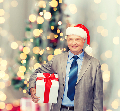 business, presents and people concept - smiling senior man in suit and santa helper hat with gift over christmas tree lights background