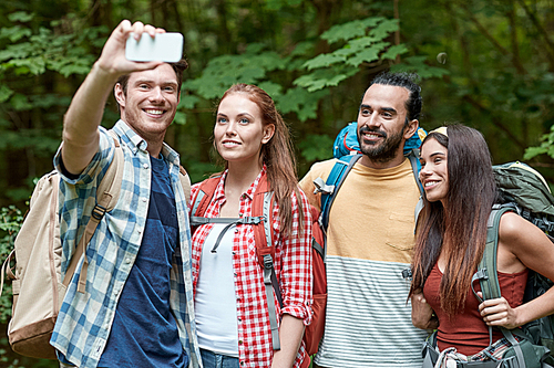 technology, travel, tourism, hike and people concept - group of smiling friends walking with backpacks taking selfie by smartphone in woods