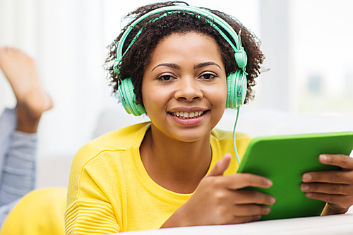 people, technology and leisure concept - happy african american young woman lying on sofa with tablet pc computer and headphones listening to music at home