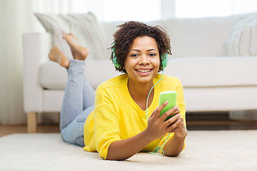 people, technology and leisure concept - happy african american young woman lying on floor with smartphone and headphones listening to music at home