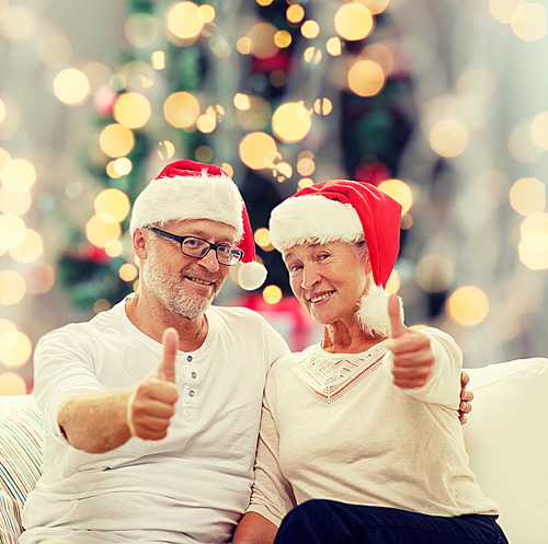 family, holidays, age and people concept - happy senior couple in santa helper hats sitting on sofa over christmas tree lights background