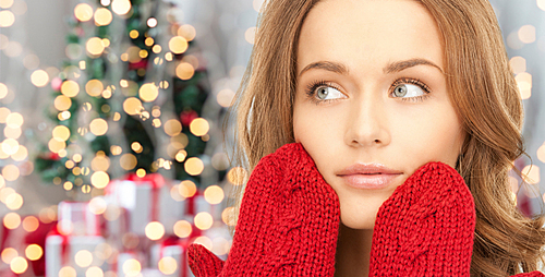 winter holidays, christmas and people concept - happy young woman in red mittens touching her face over christmas tree lights background