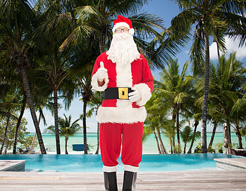 christmas, holidays, gesture, travel and people concept- man in costume of santa claus showing thumbs up over swimming pool on tropical beach background