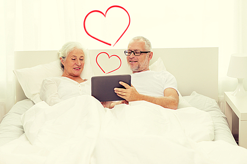 family, technology, love, age and people concept - happy senior couple looking to tablet pc computer and lying in bad at home with red heart shapes