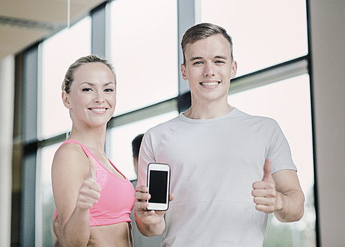 fitness, sport, advertising, technology and diet concept - smiling young woman and personal trainer with smartphone in gym showing thumbs up