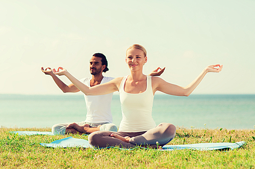 fitness, sport, friendship and lifestyle concept - smiling couple making yoga exercises sitting on mats outdoors