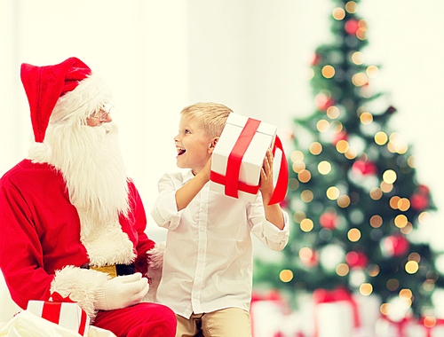 holidays, childhood and people concept - smiling little boy with santa claus and gifts over christmas tree lights background