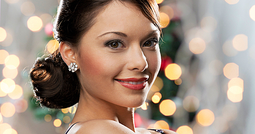 people, holidays, jewelry and luxury concept - smiling woman face with diamond earring over christmas lights background