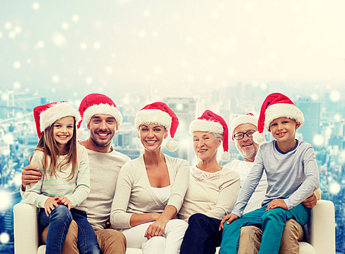 family, happiness, generation, holidays and people concept - happy family in santa helper hats sitting on couch over snowy city background