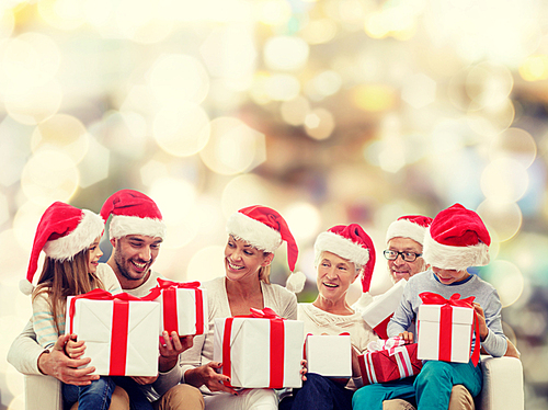 family, happiness, generation, holidays and people concept - happy family in santa helper hats with gift boxes sitting on couch over lights background