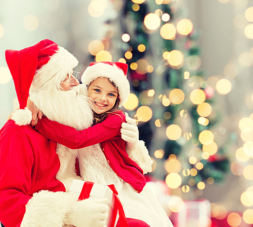 holidays, celebration, childhood and people concept - smiling little girl hugging with santa claus over christmas tree lights background