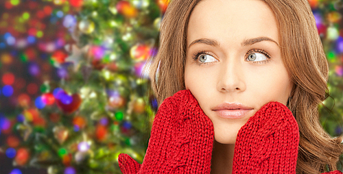 happiness, winter holidays, christmas and people concept - happy young woman in red mittens touching her face over lights background