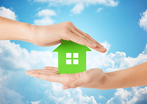 eco, people and real estate concept - close up of woman hands holding green house over blue sky and clouds background