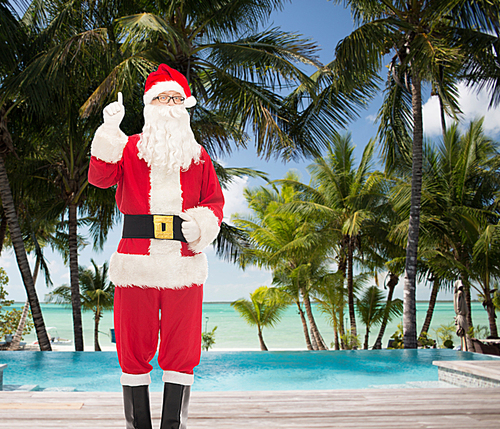 christmas, holidays, gesture, travel and people concept - man in costume of santa claus pointing finger up over tropical beach and swimming pool background