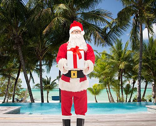 christmas, holidays, travel and people concept - man in costume of santa claus with gift box over swimming pool on tropical beach background